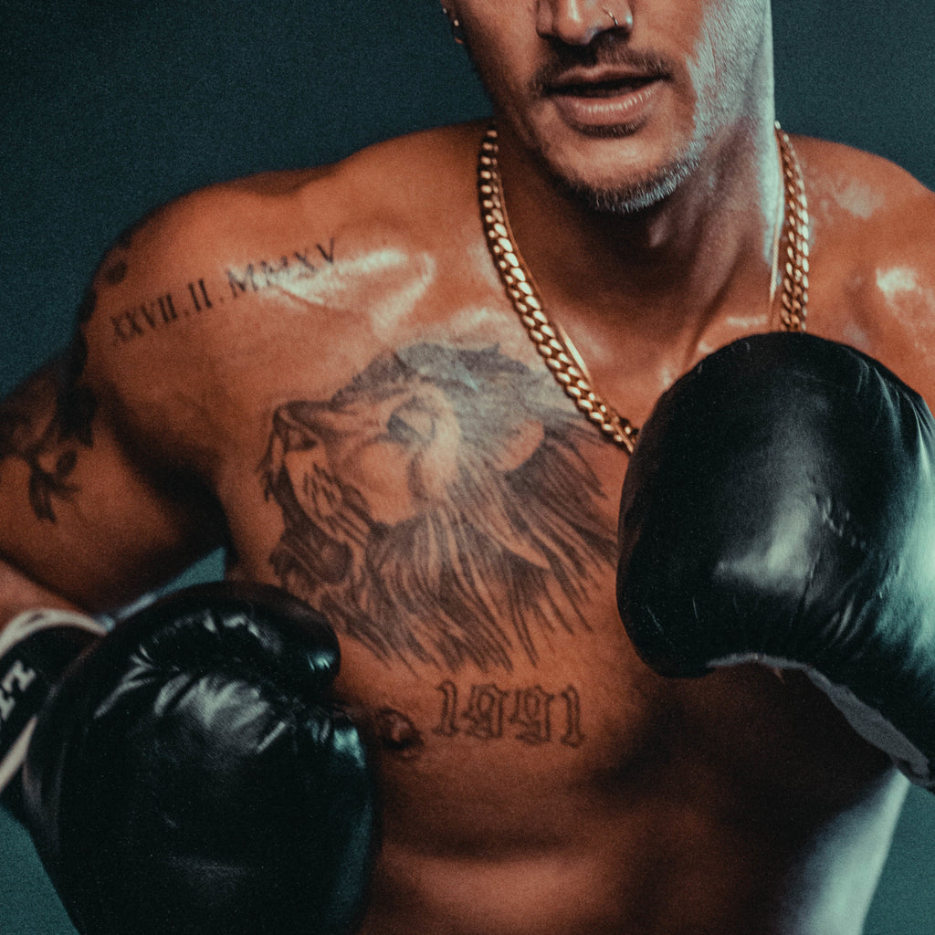 A topless man in boxing gloves. He has tattoos and is wearing a gold chain.