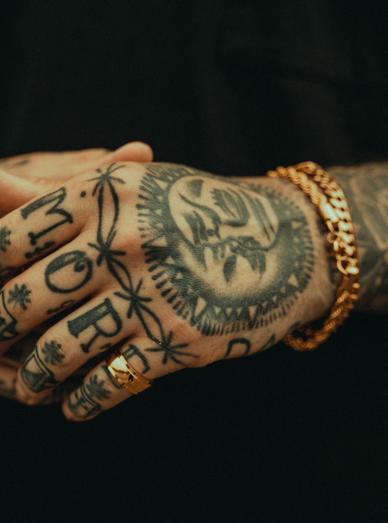 a tattooed hand in a gold ring and chain. MORE tattooed across the knuckles.