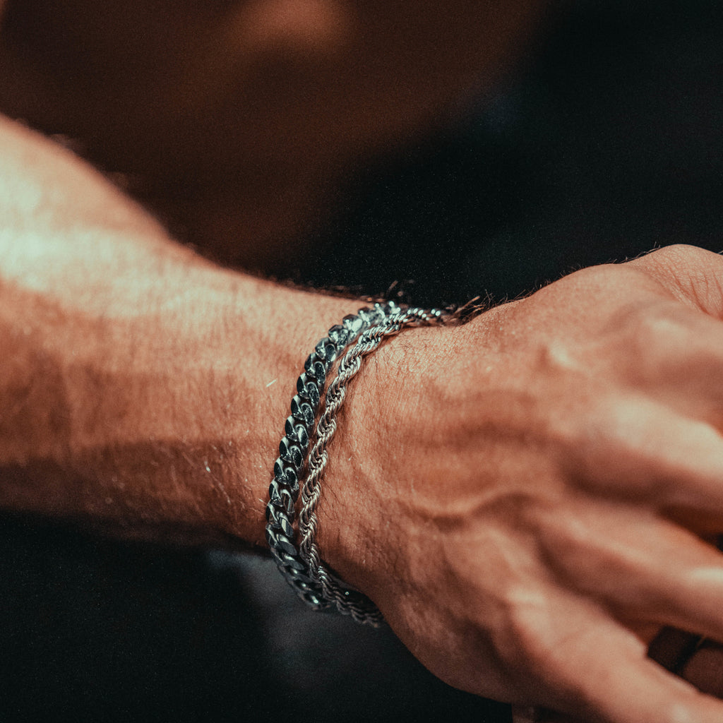 Man's hand wearing two platinum bracelets: cuban link and rope chain.